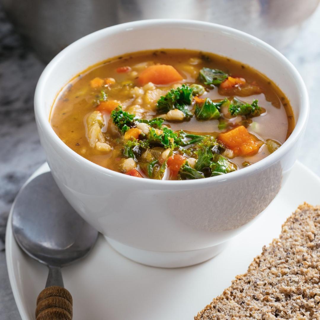 A white bowl filled with hearty vegetable soup containing kale, carrots, and tomatoes sits on a plate with a slice of whole grain bread and a spoon next to it.